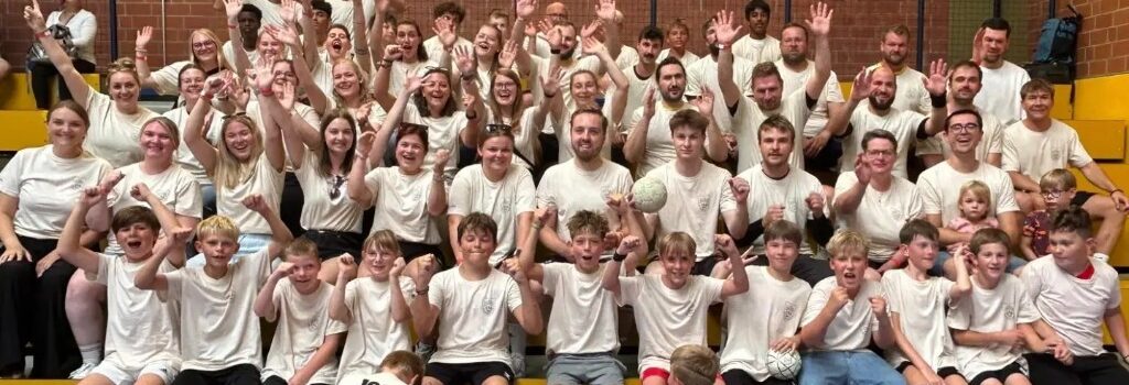 Die Handballabteilung der TSG in der Halle, viele in weißen Shirts. Zwei Kinder vorne tragen Shirts mit dem Aufdruck 100 Jahre Handball.