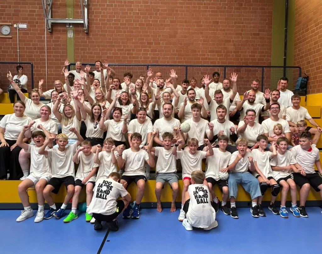 Die Handballabteilung der TSG in der Halle, viele in weißen Shirts. Zwei Kinder vorne tragen Shirts mit dem Aufdruck 100 Jahre Handball.