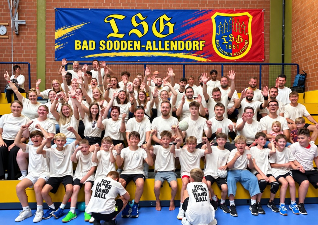 Die Handballabteilung der TSG in der Halle vor dem blau-roten Vereinsbanner. Viele tragen weiße Shirts, zwei Kinder vorne Shirts mit dem Aufdruck 100 Jahre Handball.